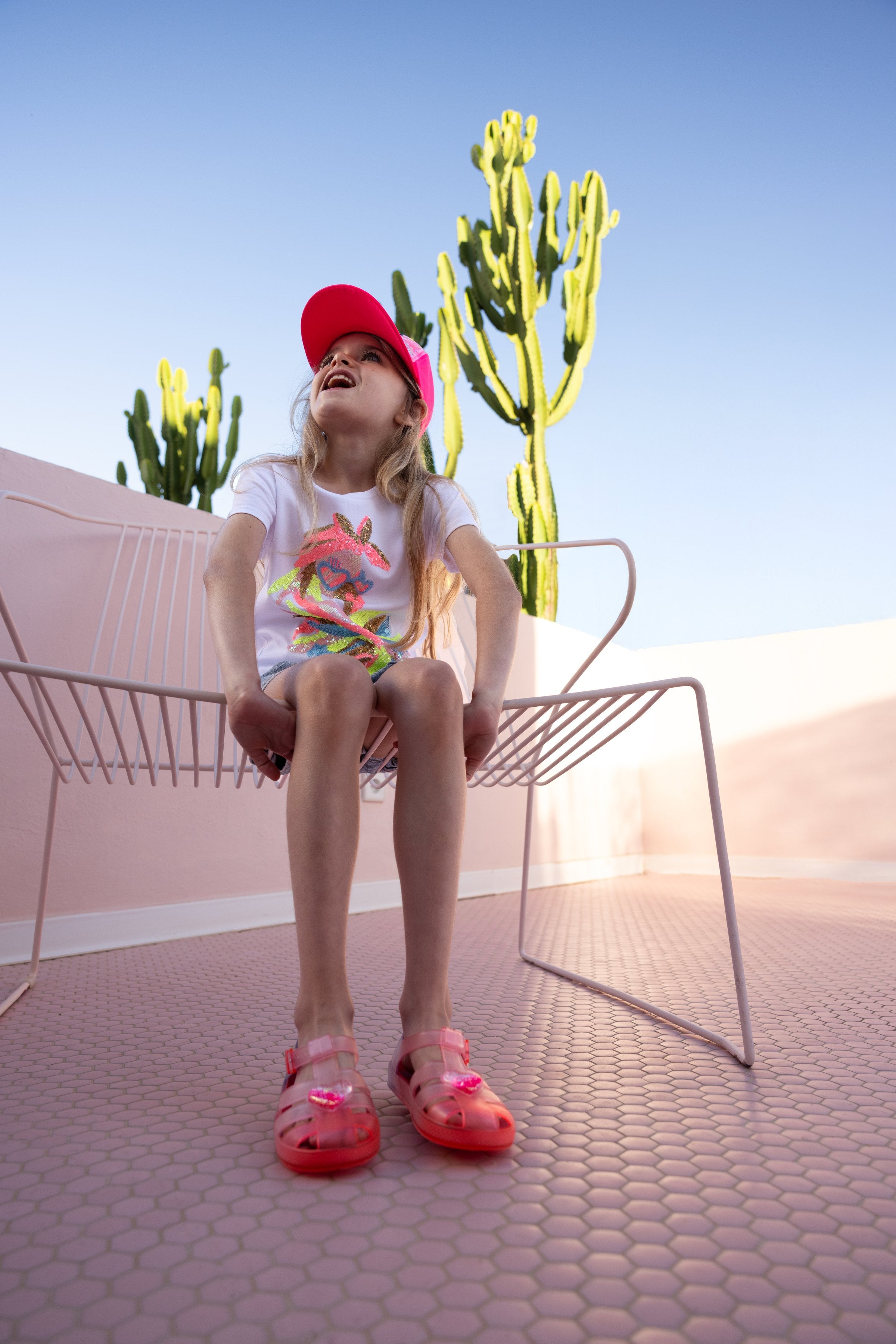 Meisje in zomerse Billieblush outfit met rode pet en roze sandalen in de zon — zomerse kindermode bij Niñoz Almere